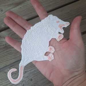 Peut inclure: Patch thermocollant brodé blanc représentant un opossum avec des détails roses. L'opossum est tourné vers la droite de l'image et a une queue enroulée.