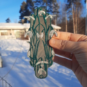 以下が含まれることがあります： 白い花と緑の葉が刺繍されたパッチ。雪割草の花が密集して描かれている。パッチは透明な背景に置かれている。
