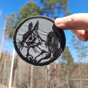 May include: A gray round patch with a black embroidered image of a squirrel. The squirrel is facing left and has its tail curled around its body.