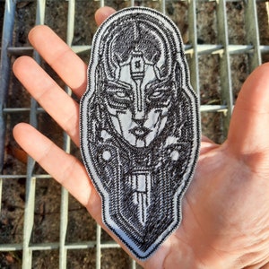 May include: A black and white embroidered patch depicting a detailed illustration of a futuristic, humanoid face with a helmet-like headpiece. The face has a serious expression and is surrounded by intricate details.