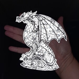 Puede incluir: Un parche de hierro reflectante plateado de un dragón con las alas extendidas. El dragón está en posición sentada con la cabeza girada hacia un lado. El parche tiene un diseño detallado con escamas y una superficie texturizada.