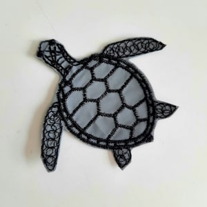 Op de afbeelding: Een grijze stoffen zeeschildpad patch met zwarte geborduurde details. De schildpad heeft een gedetailleerde schild en flippers.
