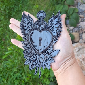 Può includere: Una toppa grigia a forma di cuore con un design a buco della serratura, circondata da rose nere, ali e un lucchetto. La toppa è tenuta in una mano, mettendo in risalto i dettagli del ricamo. È un oggetto decorativo.