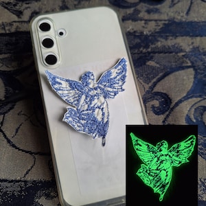 Könnte beinhalten: Eine weiße Smartphone-Hülle mit einem gestickten Aufnäher eines Engels mit ausgebreiteten Flügeln. Die Stickerei ist bei Tageslicht blau und leuchtet im Dunkeln hellgrün, wie im kleinen Bild in der unteren rechten Ecke zu sehen ist.