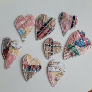 May include: A collection of eight handmade heart-shaped fabric patches. Each heart features a unique patchwork design with various patterns and colors, including pink, beige, and blue. The edges are finished with decorative stitching and some have small bead embellishments.