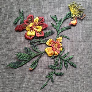 May include: An embroidered floral patch featuring two large blossoms with deep red and bright yellow petals, a smaller yellow bud at the top, and detailed green stems with serrated leaves. The embroidery uses dense thread work to create texture.