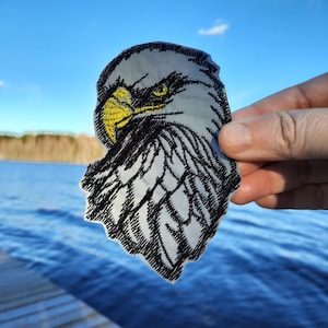 Op de afbeelding: Close-up van een geborduurde patch van een Amerikaanse zeearendkop met een gele snavel en zwarte ogen. De adelaar kijkt naar links en de achtergrond is wit.