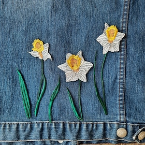 Könnte beinhalten: Drei gestickte weiße Narzissen mit gelben Zentren sind auf eine blaue Jeansjacke genäht. Die Blumen haben grüne Stiele und Blätter.