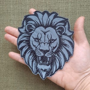 Op de afbeelding: Een grijs geborduurd pleister met een leeuwenkop van zwart garen. De leeuw heeft een woeste uitdrukking met zijn bek open en zichtbare tanden.