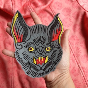 May include: A gray embroidered iron-on patch of a bat with red and yellow details. The bat has sharp teeth and large eyes.