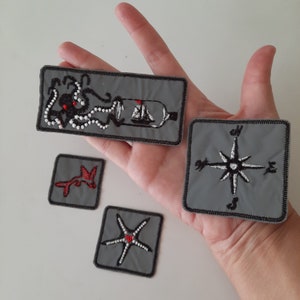 May include: Four embroidered iron-on patches with nautical themes. The patches are grey with black and white embroidery. One patch features an octopus with a bottle and a ship inside. Another patch features a compass with the directions North, South, East, and West. The third patch features a starfish. The fourth patch features a red and white aeroplane.