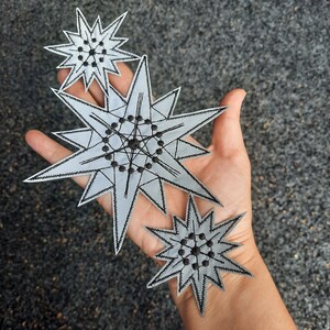Könnte beinhalten: Drei silberne sternförmige Patches mit schwarzen Nähten und einem geometrischen Design. Die Sterne sind übereinander gelegt.