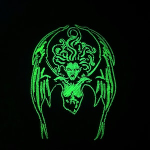 May include: A green glow-in-the-dark embroidered patch featuring a winged female figure with snakes for hair. The figure has a fierce expression and is depicted with wings outstretched.