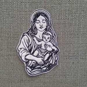May include: Black and white embroidered patch of the Virgin Mary holding a baby Jesus. The patch is on a textured fabric background.