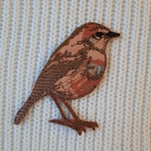 以下が含まれることがあります： 茶色とピンクの刺繍でできた鳥のアップリケで、細部まで丁寧にデザインされています。鳥は白いニットの表面に置かれています。