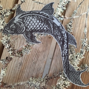 Könnte beinhalten: Ein grauer und schwarzer Fisch-Patch mit einem detaillierten Design. Der Patch ist aus Stoff und hat eine raue Textur.