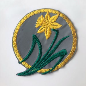 May include: A round, gray embroidered patch with a yellow daffodil and green leaves. The patch has a gold border.