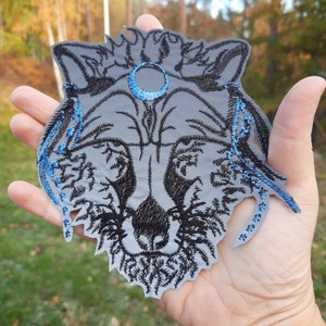 May include: A gray embroidered patch of a wolf's face with a blue moon above it. The wolf's fur is detailed with black embroidery and the moon is outlined in blue thread.