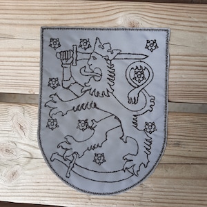 Puede incluir: Un parche bordado gris que presenta el escudo de armas de Finlandia. El parche representa un león sosteniendo una espada, rodeado de flores estilizadas.