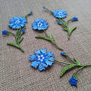 Könnte beinhalten: Fünf blaue gestickte Blumenapplikationen mit grünen Stielen und Blättern. Die Blüten haben weiße Zentren und sind auf einem braunen Leinenstoff angeordnet.