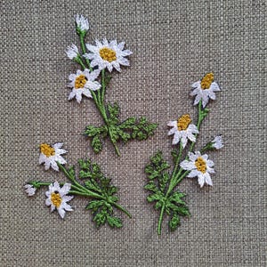 Puede incluir: Cuatro parches bordados de margaritas blancas con tallos y hojas verdes sobre un fondo de tela marrón.