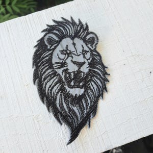 May include: Black and white embroidered lion head patch. The lion has a fierce expression with its mouth open and teeth showing. The patch is perfect for adding a touch of wildness to any project.