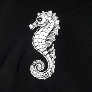 Könnte beinhalten: Ein weiß gesticktes Seepferdchen-Design auf einem schwarzen Stoffhintergrund.