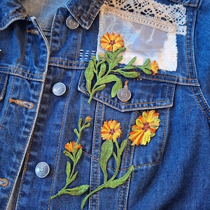 Puede incluir: Una chaqueta vaquera azul con flores bordadas en naranja y amarillo y hojas verdes. La chaqueta tiene botones plateados y un parche de encaje blanco y tela en la parte superior derecha.