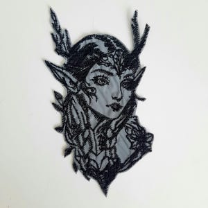 May include: Black and white embroidered patch of a woman with antlers and a floral design. The woman has long hair and is wearing a dress with a floral pattern.