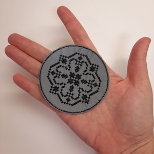 May include: A gray circular fabric patch with a black embroidered geometric design. The design resembles a stylized snowflake or star.