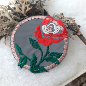 May include: A round embroidered patch featuring a red rose with white and pink accents on a grey background. The patch has a pink border.