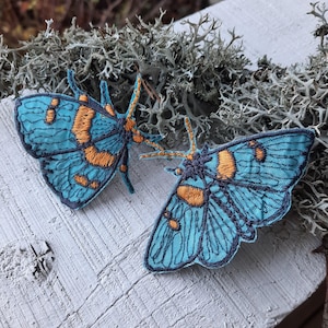 以下が含まれることがあります： 黒い輪郭のある、青とオレンジの刺繍が施された蛾のアップリケが2つ。蛾は白木の表面に置かれ、薄い灰色の背景がある。