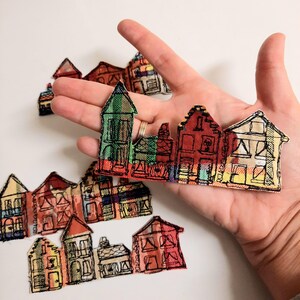 Può includere: Un set di applicazioni in tessuto colorate che raffigurano una fila di case. Ogni casa è delineata in nero e ha un design e uno schema di colori unici. Le applicazioni sono perfette per aggiungere un tocco fantasioso a qualsiasi progetto artigianale.