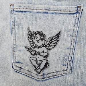 May include: A light blue denim jean pocket with a black and white embroidered patch of a cherub with wings.