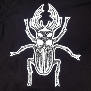 Könnte beinhalten: Ein weißes Hirschkäfer-Design auf einem schwarzen Stoffhintergrund. Der Käfer ist mit filigranen Linien und Schattierungen detailliert.