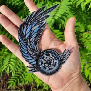 May include: A black and blue embroidered patch with a wing design and a circular emblem in the center. The emblem has a black and white design with a star in the center.