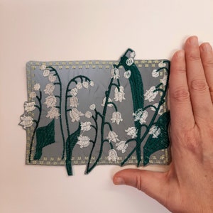 May include: A grey fabric patch with an embroidered design of white lily of the valley flowers with green leaves. The patch has a decorative stitched border.
