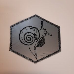 May include: A silver hexagonal patch with a black embroidered snail design.