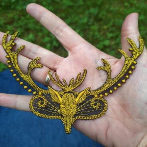 Può includere: Toppa ricamata raffigurante una testa di cervo e corna dorate su sfondo marrone scuro. La toppa presenta dettagli intricati e un bordo decorativo. Le corna sono ornate da piccole perline dorate.