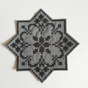 Pode incluir: Um descanso de copo em forma de estrela de tecido cinzento com um padrão geométrico bordado preto.