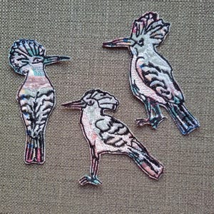 May include: Three embroidered iron-on patches of hoopoes, a type of bird, in shades of pink, blue, and white. The patches are on a brown fabric background.