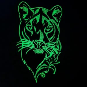 Puede incluir: Un parche bordado verde que brilla en la oscuridad con la cara de un puma y un diseño floral.