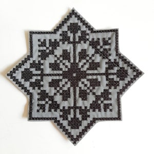 May include: A gray, eight-pointed star-shaped textile with a detailed black cross-stitch pattern. The design features a central motif and repeating geometric elements, creating a symmetrical and intricate appearance. The piece is likely handmade.