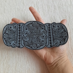 May include: A gray fabric patch with a black embroidered design of an intricate knot pattern. The patch is rectangular and has a slightly rounded edge.