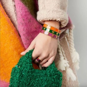 Puede incluir: Un conjunto de cuatro pulseras de brazalete doradas con esmalte en tonos de morado, verde y naranja. Cada pulsera está decorada con pequeños corazones dorados.