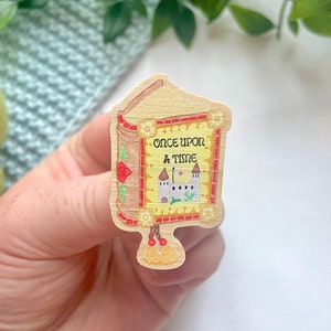 May include: Wooden pin shaped like a storybook with the words "ONCE UPON A TIME" and a castle illustration. The pin has a light brown background with red and yellow accents, and a tassel at the bottom.