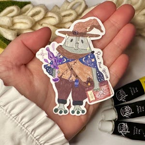Könnte beinhalten: Ein skurriler Aufkleber mit einem Cartoon-Zaubererfrosch mit braunem Hut und blauem Sternenumhang. Der Aufkleber trägt den Text "Pick Your Poison". Der Frosch hält einen lila Trank und ein kleines weißes Kaninchen. Acrylfarbtuben sind im Hintergrund zu sehen.