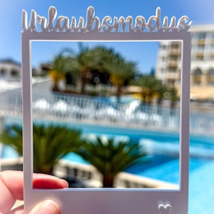 May include: A white, Polaroid-style frame with the German text "Urlaubsmodus" at the top and a small heart at the bottom right. The frame is held up against a blurred background of a pool, palm trees, and a building under a bright blue sky.