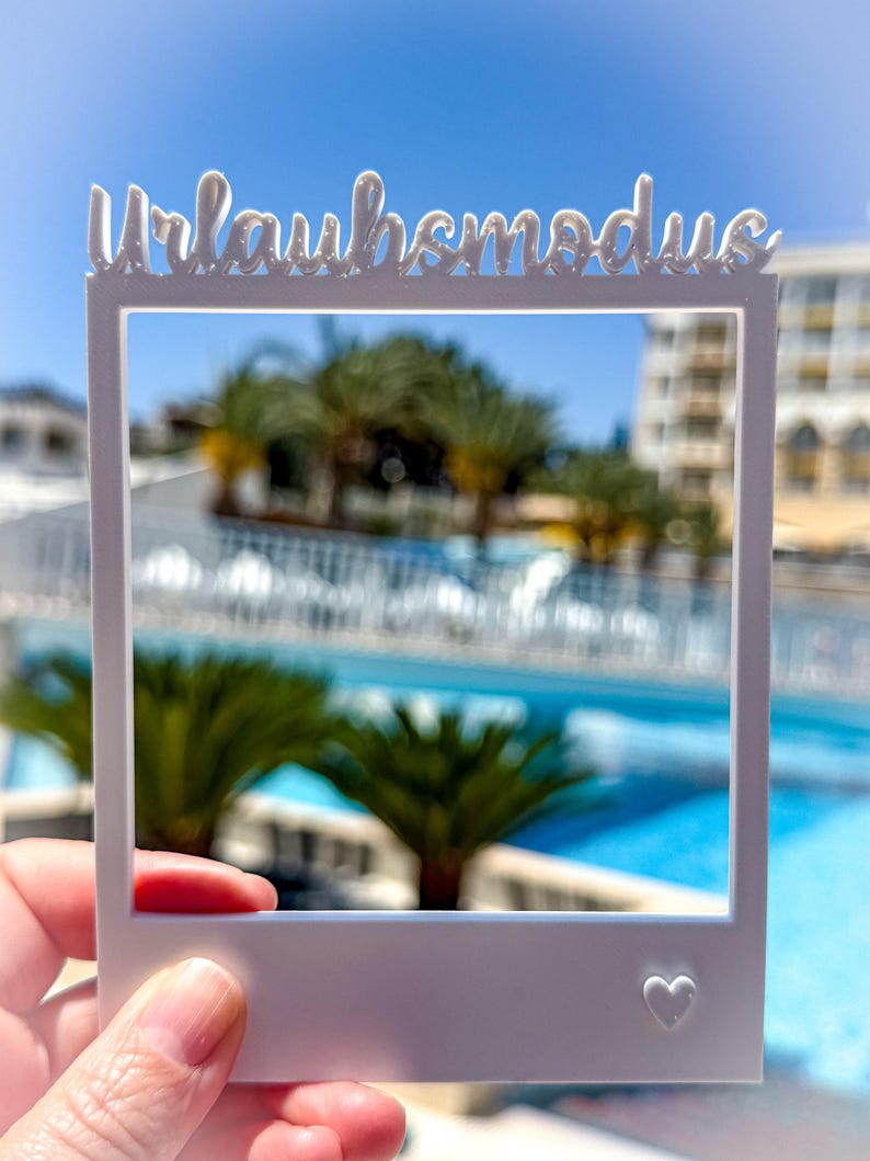 May include: A white, Polaroid-style frame with the German text "Urlaubsmodus" at the top and a small heart at the bottom right. The frame is held up against a blurred background of a pool, palm trees, and a building under a bright blue sky.