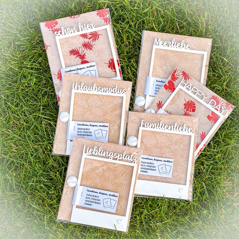 May include: A collection of framed cards with various German phrases in white script, including "schon hier" and "Happy Day." The cards have a beige background with red leaf patterns and are arranged on green grass.